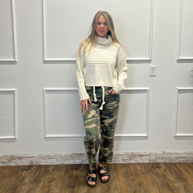 A woman stands in front of a white paneled wall wearing a cream ribbed turtleneck sweater and camouflage drawstring pants, paired with black sandals.
