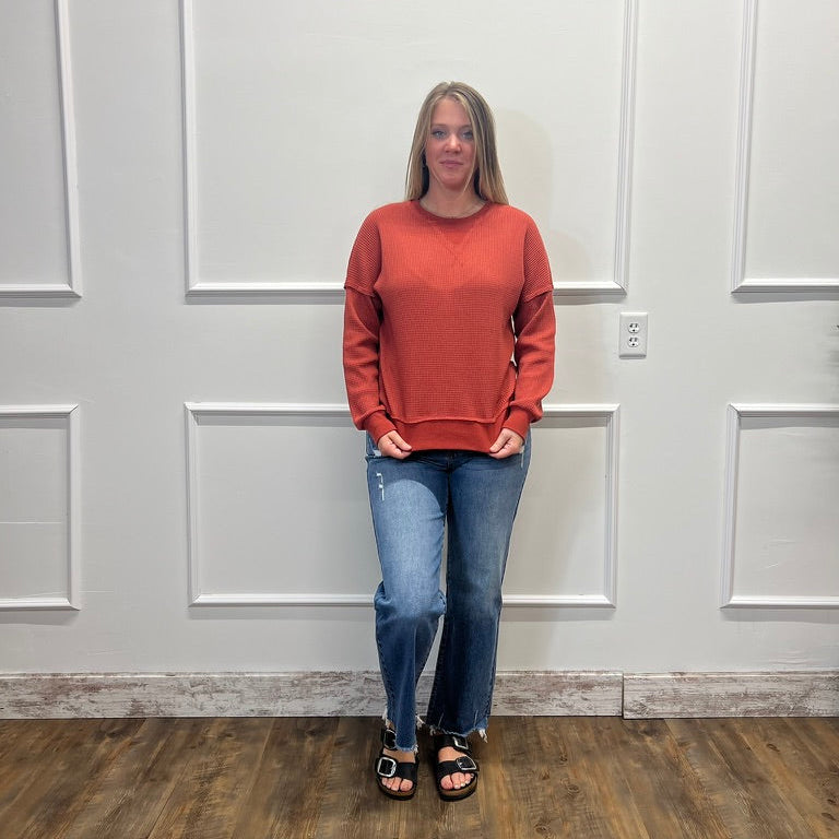 A woman stands in a room with white paneled walls and a wood floor, wearing a rust-orange long-sleeve crewneck sweatshirt, blue jeans, and black sandals.