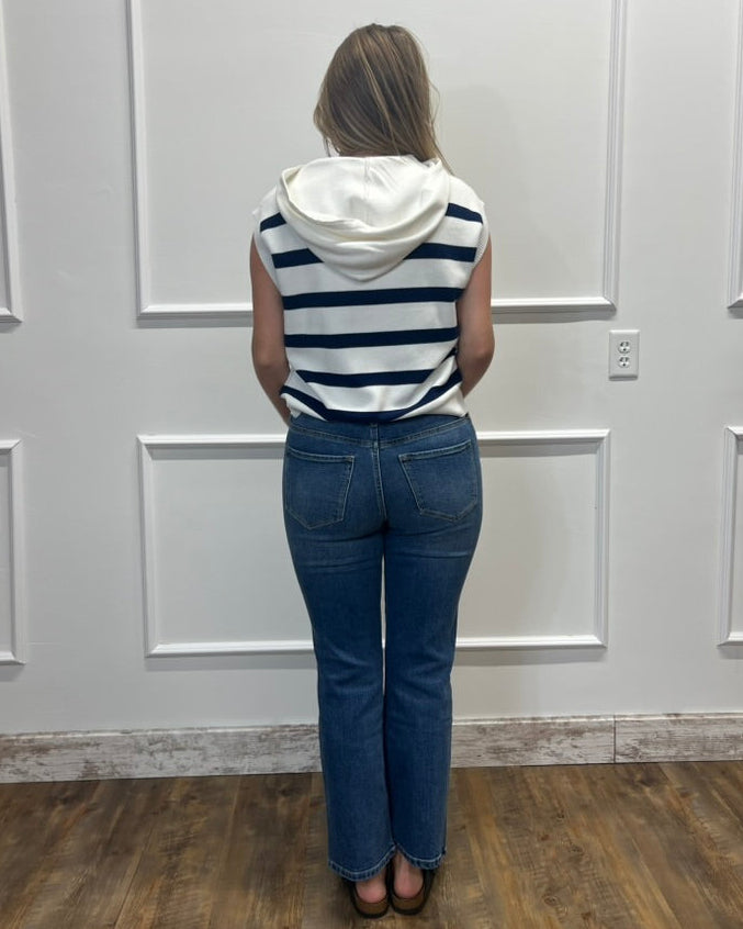 A woman viewed from behind wearing a sleeveless navy and white striped hoodie top with blue jeans, standing in front of a white paneled wall.