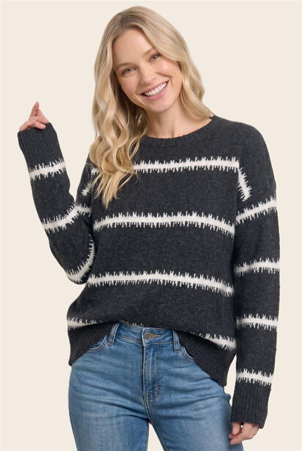 A smiling blonde woman wearing a dark charcoal gray knit sweater with horizontal white textured stripes and blue jeans, posed against a solid light beige background.