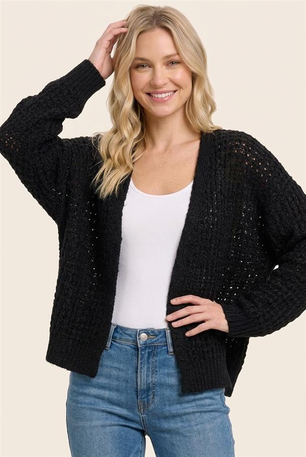 A woman wearing a black open-front knit cardigan with textured openwork details, styled over a white tank top and blue jeans, against a solid light beige background.