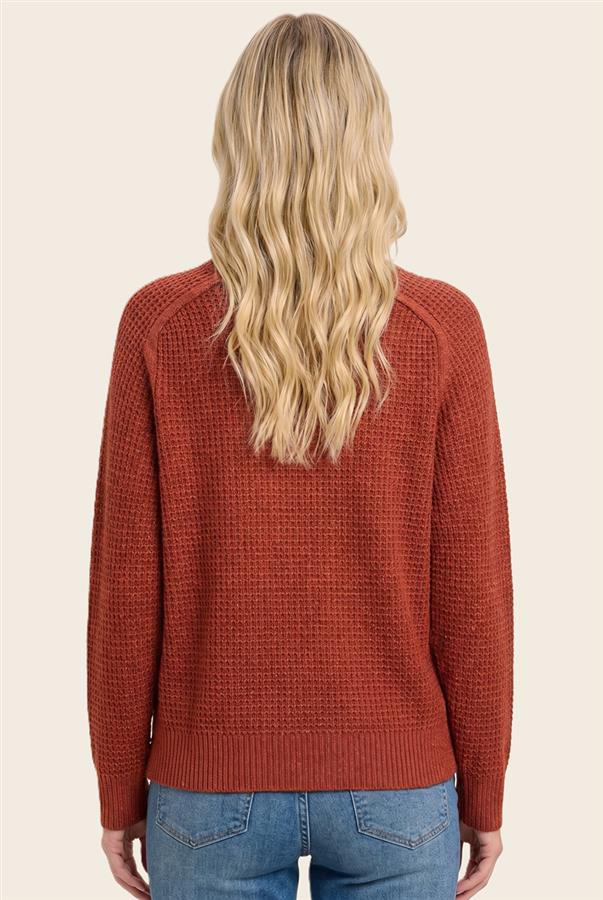 A woman viewed from the back wearing a rust-orange textured knit sweater and blue jeans against a plain light beige background.