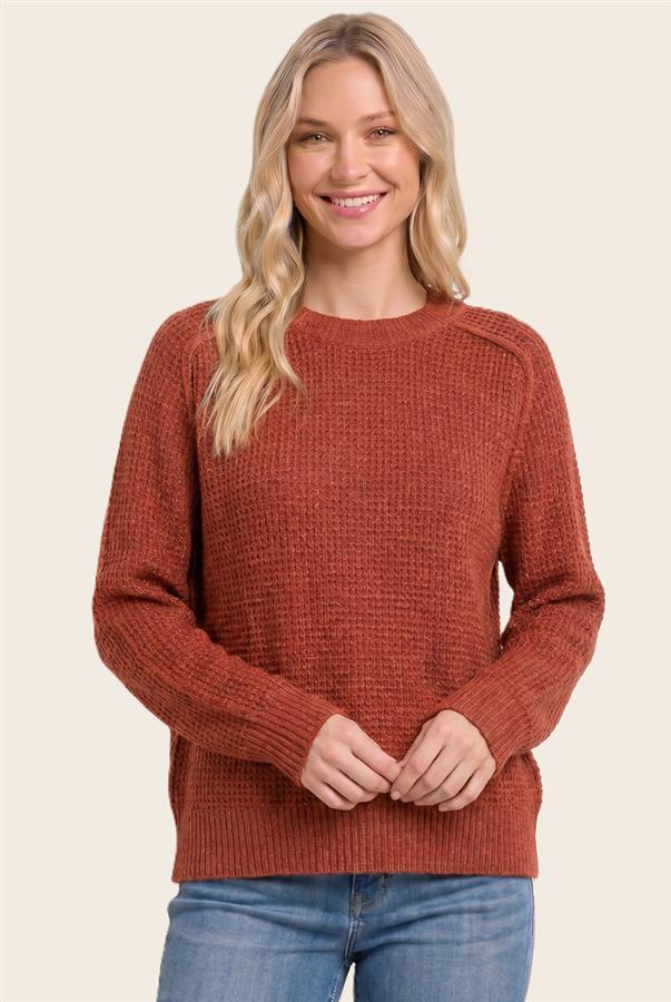 A woman wearing a textured rust-orange crewneck sweater and blue jeans, standing against a solid light beige background.