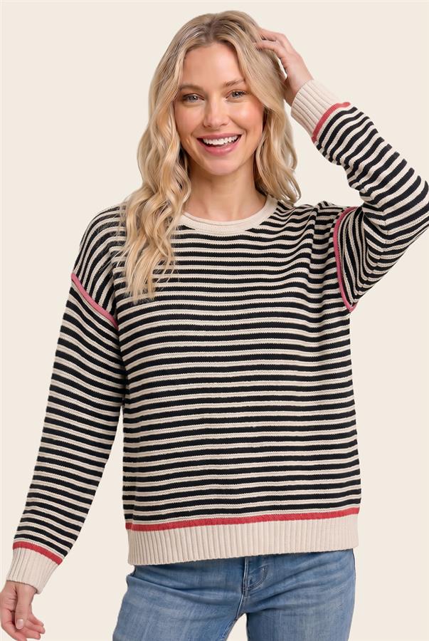 A woman wearing a black and cream striped knit sweater with thin red stripes at the cuffs and hem, paired with blue jeans, against a solid light beige background.