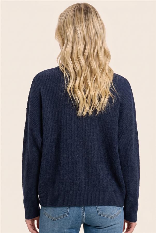 A woman viewed from the back wearing a navy blue ribbed knit sweater and light blue jeans against a plain beige background.