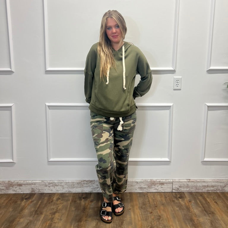 A woman stands indoors against a white paneled wall wearing an olive green hoodie with white drawstrings and camouflage jogger pants, paired with black sandals.