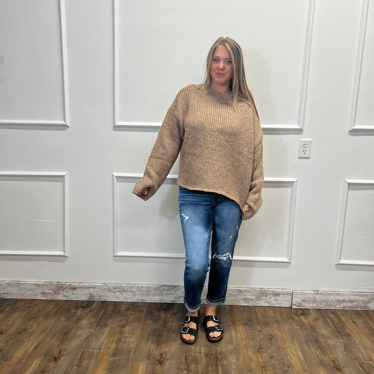 A woman modeling a beige ribbed knit mock neck sweater, paired with blue jeans and black sandals, standing in front of a white paneled wall on a wood floor.