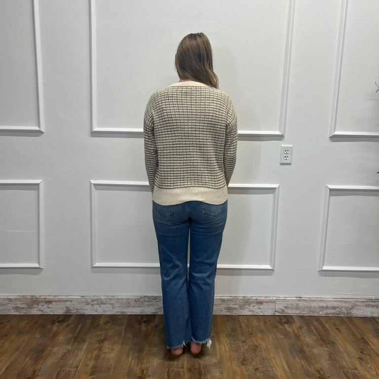 A woman stands with her back to the camera wearing a beige and black patterned knit sweater and blue jeans, standing in front of a white paneled wall on a wood floor.