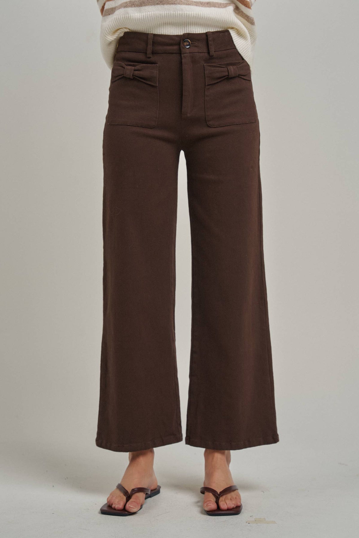 A person wearing dark brown wide-leg cropped pants with a cream and brown striped sweater and dark brown flip-flop sandals, standing against a light gray studio background.