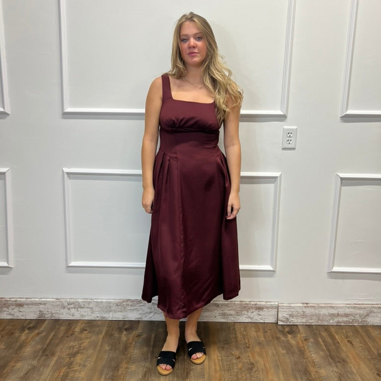 A woman modeling a sleeveless, square-neck midi dress in a deep burgundy or maroon satin-like fabric, standing against a white paneled wall on a wood floor.