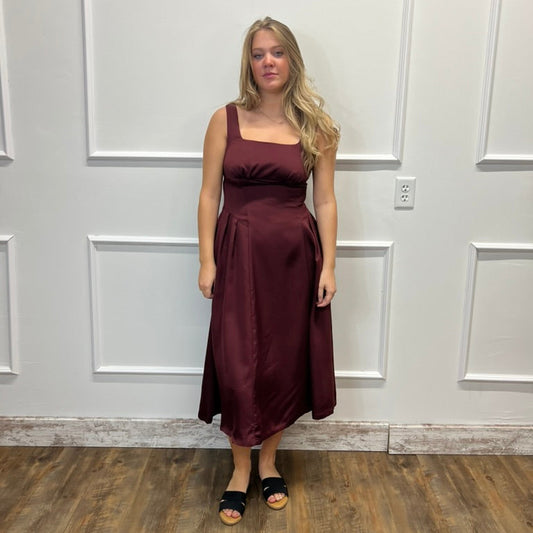 A woman modeling a sleeveless, square-neck midi dress in a deep burgundy or maroon satin-like fabric, standing against a white paneled wall on a wood floor.