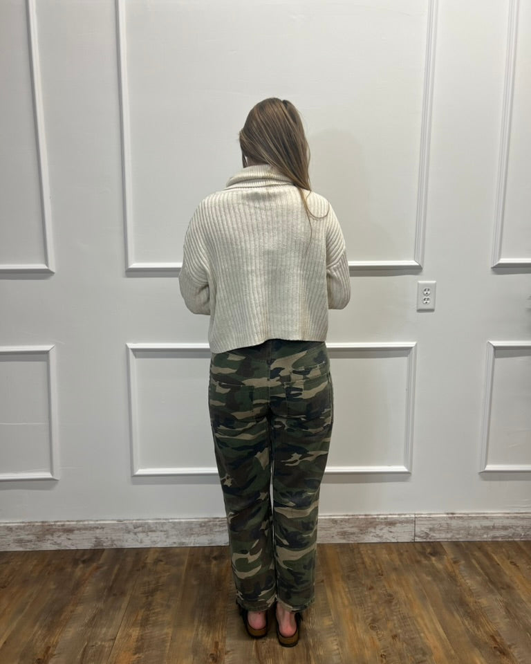 A woman viewed from behind wearing a cream ribbed knit cropped sweater and green camouflage pants, standing in front of a white paneled wall on a wood floor.