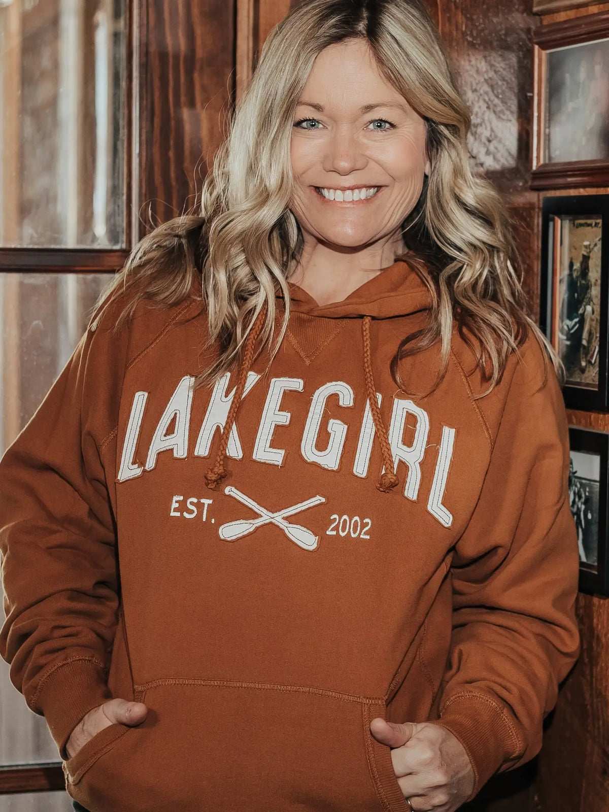 A smiling blonde woman wearing a rust-orange pullover hoodie with 'LAKEGIRL EST. 2002' and crossed oars printed in white on the front, standing indoors against a dark wood-paneled background.