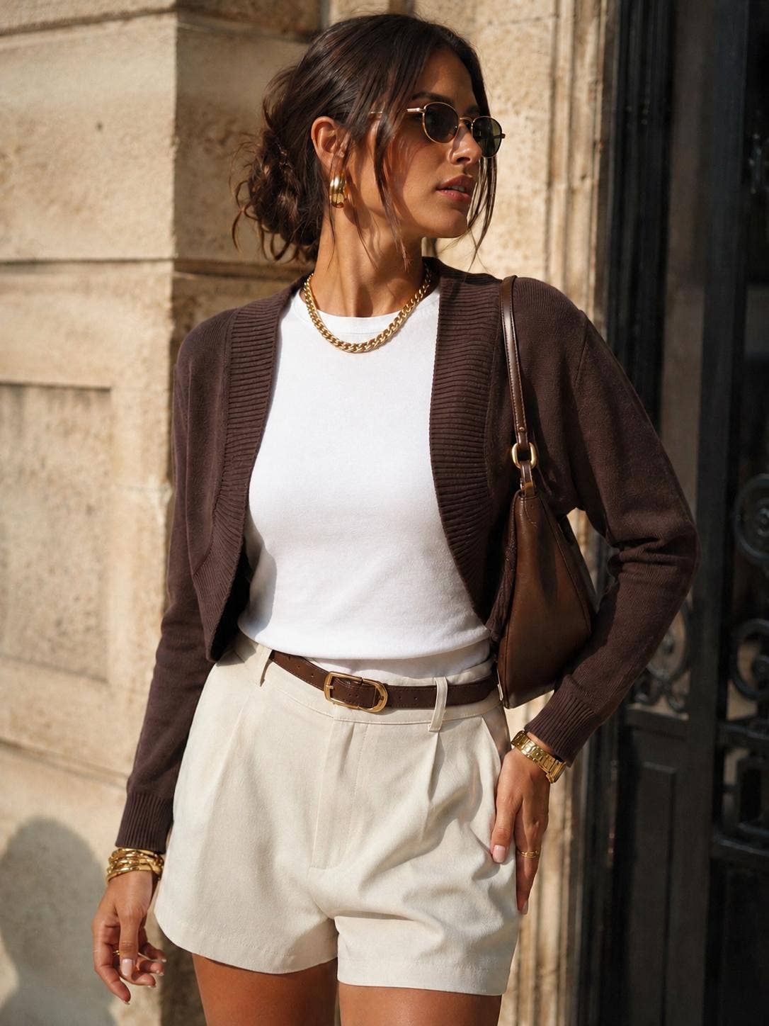 A woman wearing a dark brown ribbed cardigan over a white top, cream shorts, and gold accessories, standing outdoors against a stone wall.