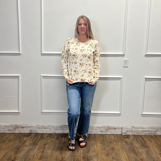 A woman stands in a room with white paneled walls and wood-look flooring, wearing a cream long-sleeve blouse with a small brown floral pattern, blue jeans, and black sandals.