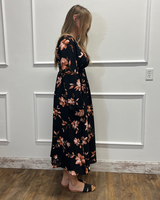 A woman wearing a black maxi dress with a pink and orange floral pattern, standing in profile against a white paneled wall on a wood floor.