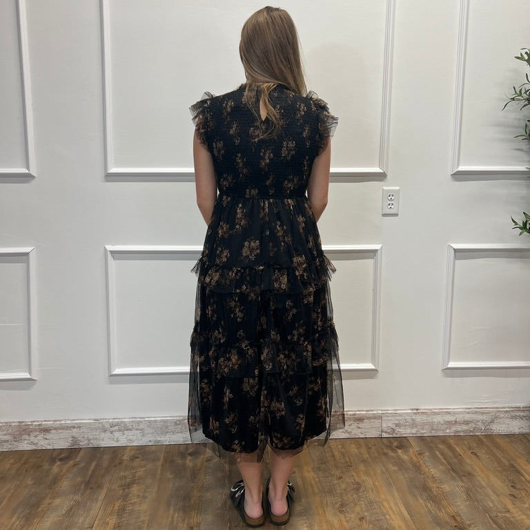 A woman stands with her back to the camera wearing a sleeveless black midi dress with a brown floral pattern and sheer tulle ruffle details. She is standing in front of a white paneled wall on a wooden floor.