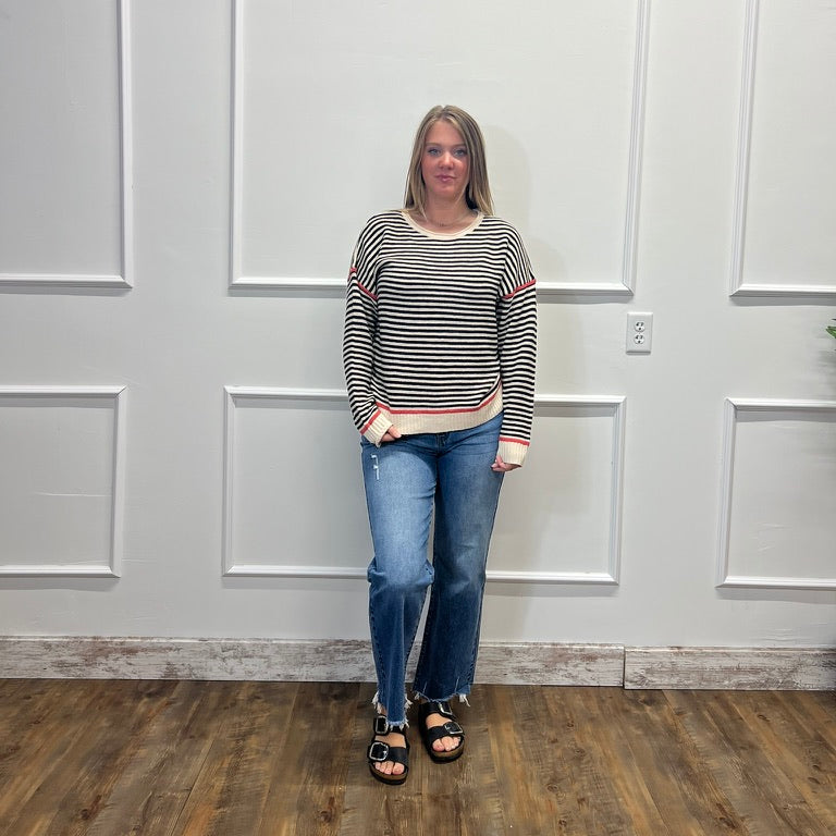 A woman stands in a room with white paneled walls and a wood floor, wearing a black and white striped long-sleeve top, blue jeans, and black sandals.