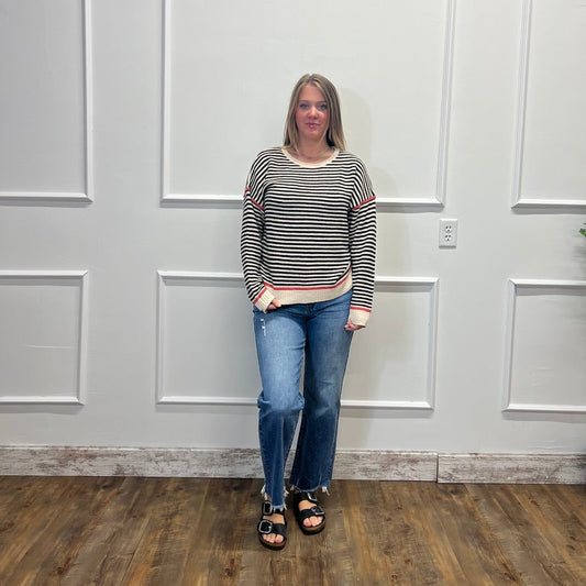 A woman stands in a room with white paneled walls and a wood floor, wearing a black and white striped long-sleeve top, blue jeans, and black sandals.