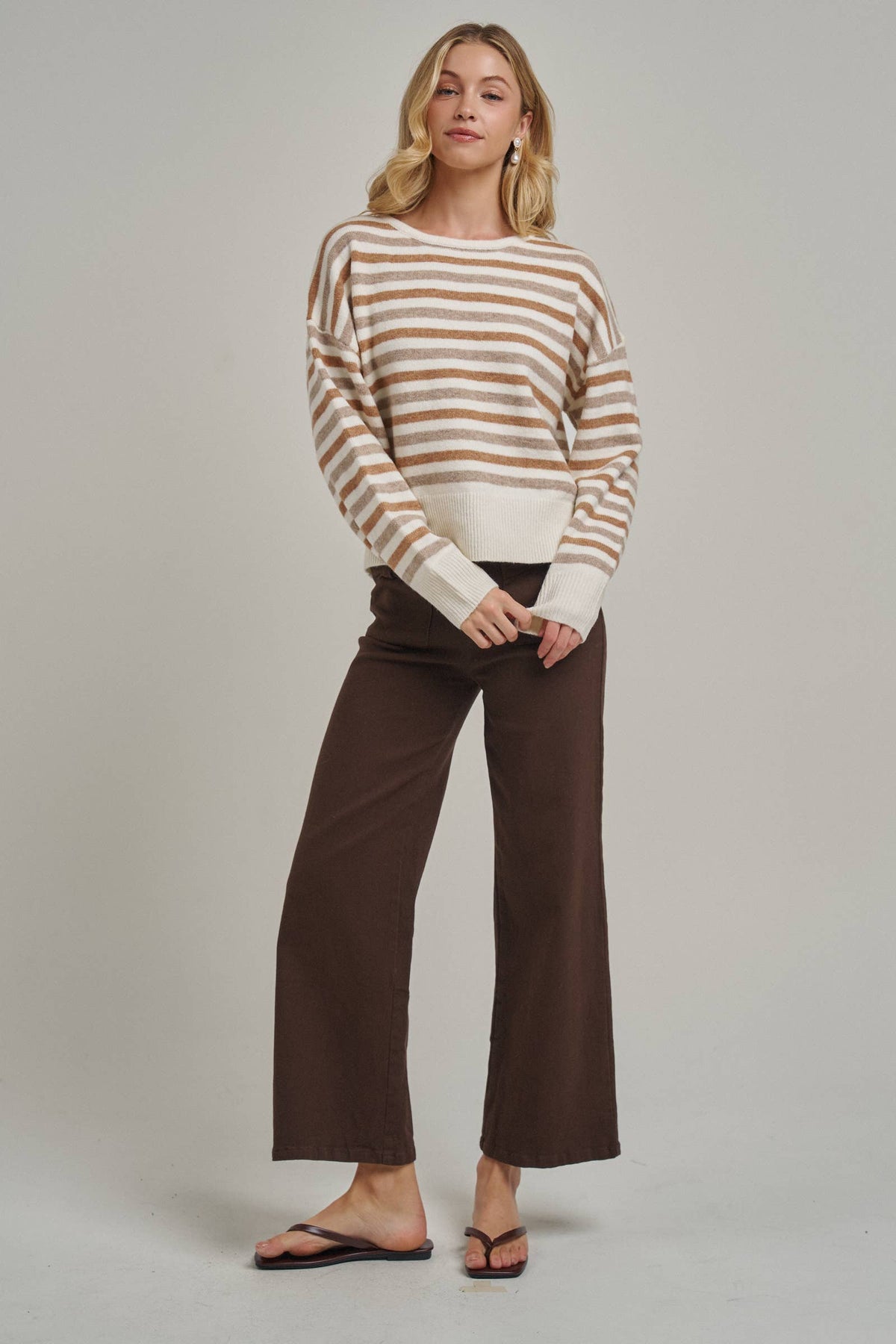 A woman modeling a long-sleeve knit sweater with horizontal cream and tan stripes, paired with dark brown wide-leg cropped pants and black flip-flop sandals, against a solid light gray studio background.