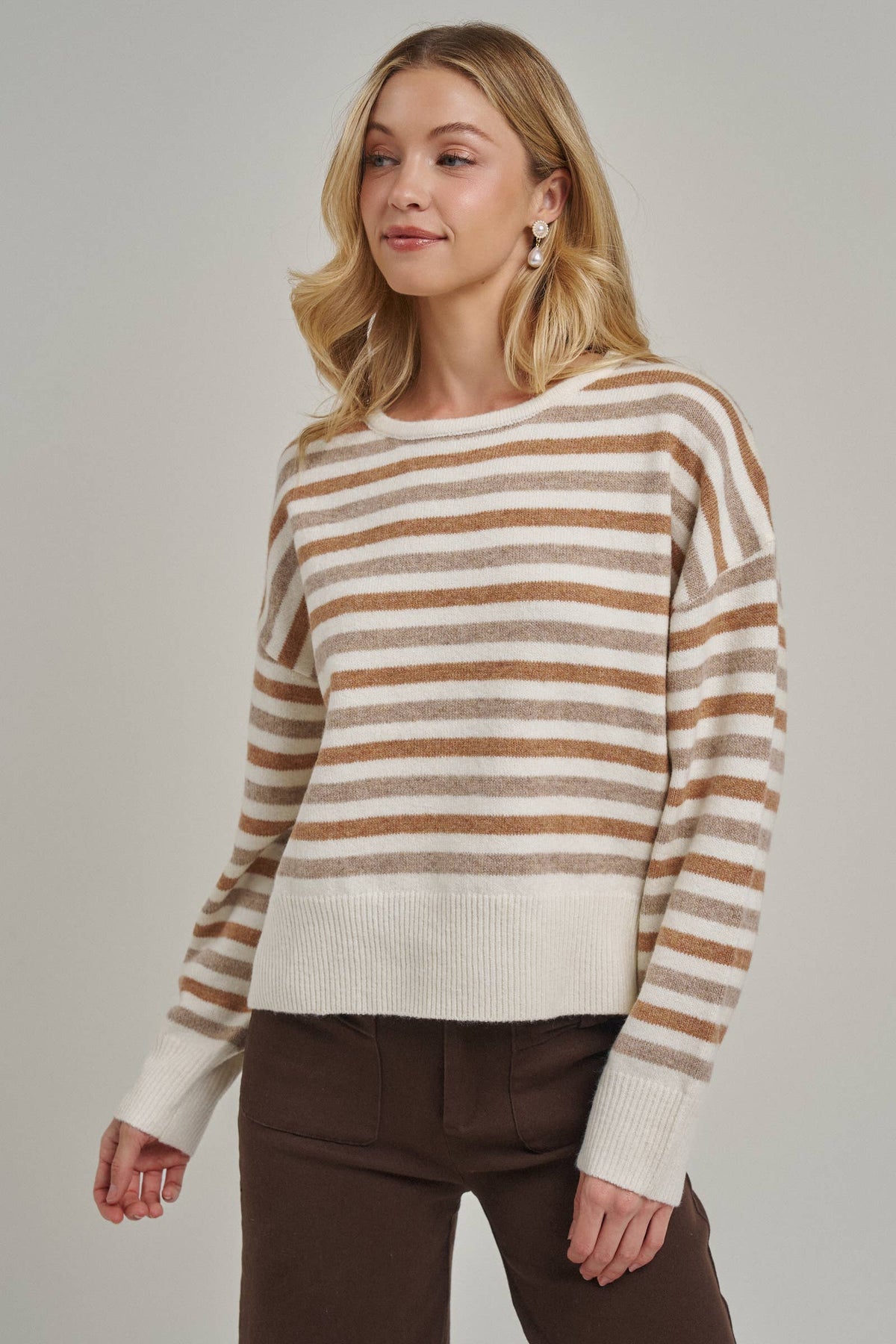 A woman wearing a cream and brown horizontally striped knit sweater with dark brown pants, photographed against a plain light gray studio background.