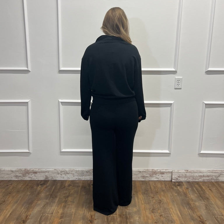 A woman stands with her back to the camera wearing a black long-sleeve top and black wide-leg pants, positioned in front of a white paneled wall on a wood-look floor.