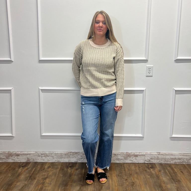 A woman stands in a room with white paneled walls and a wood floor, wearing a beige and gray textured knit sweater, blue jeans, and black slide sandals.