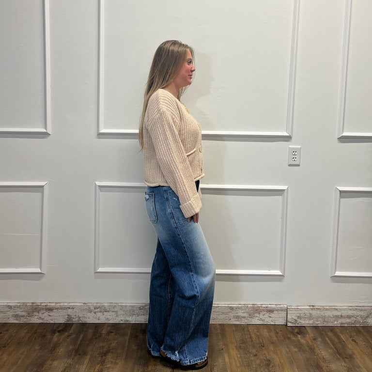 A woman stands in profile wearing a cream ribbed knit cropped cardigan and wide-leg blue jeans against a white paneled wall with wood flooring.