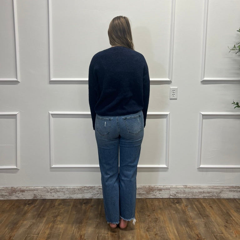 A woman viewed from behind wearing a dark navy long-sleeve top and blue jeans, standing on a wood floor against a white paneled wall.
