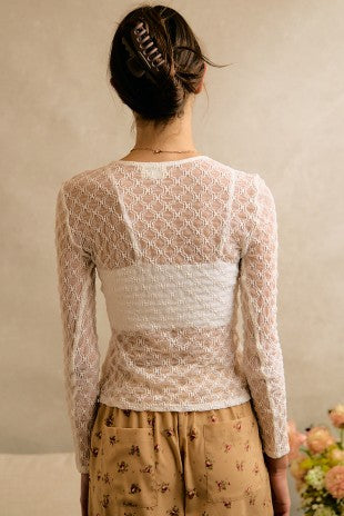 A woman shown from the back wearing a cream-colored lace long-sleeve top with a textured horizontal band across the back, paired with a tan floral skirt. She has her hair in a bun with a claw clip.