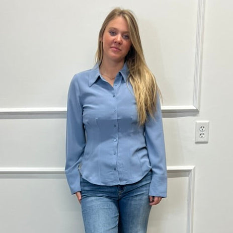 A woman stands in a room with white paneled walls and a wood floor, wearing a light blue long-sleeve button-up shirt and blue jeans.