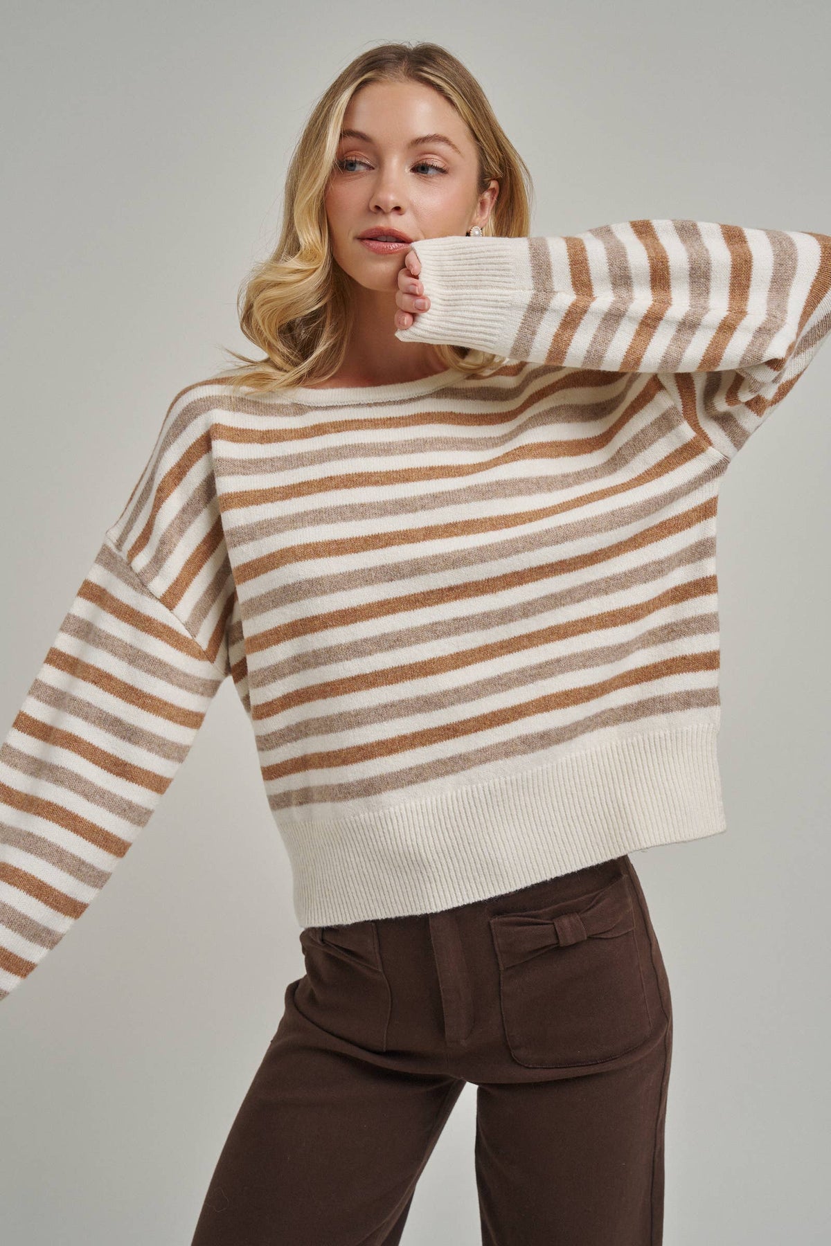 A woman wearing a cropped knit sweater with horizontal brown and cream stripes, paired with dark brown pants, against a solid light gray background.