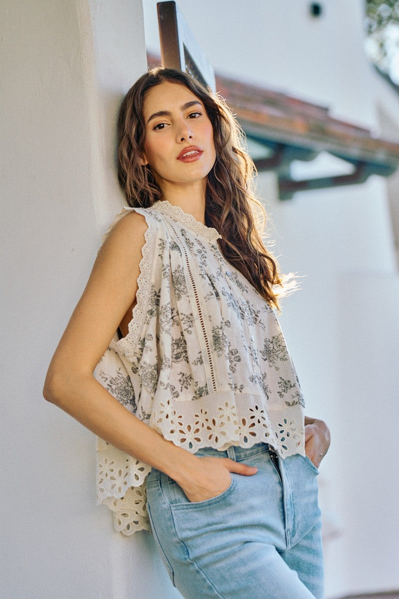 Woman wearing a floral sleeveless top and light blue jeans leaning against a white wall.