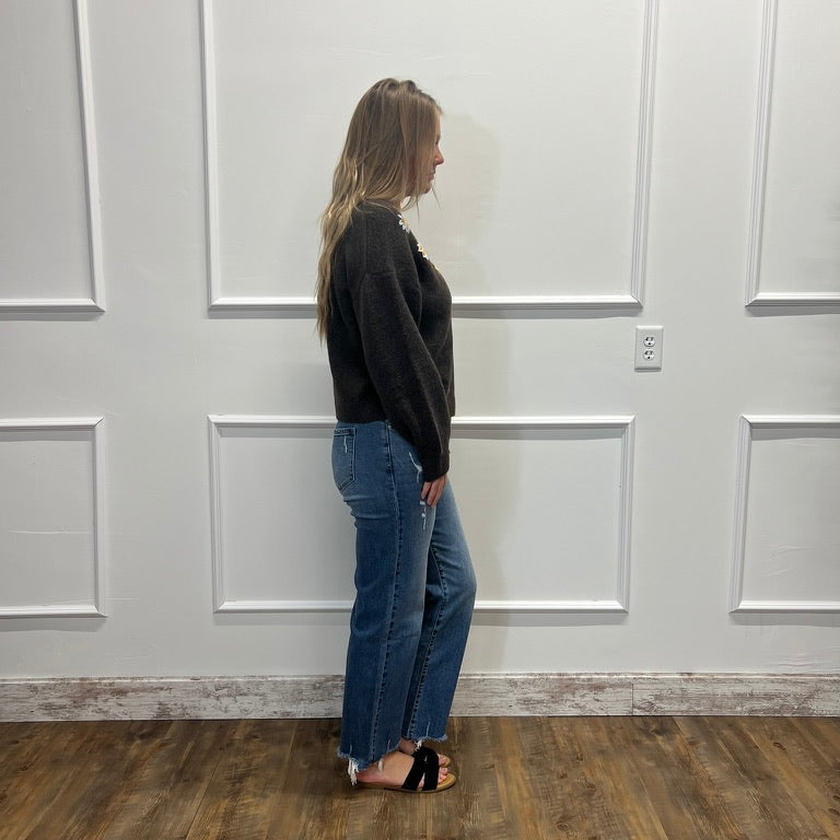 A woman stands in profile wearing a black long-sleeve top and blue wide-leg jeans with black sandals, standing on a wood-look floor against a white paneled wall.