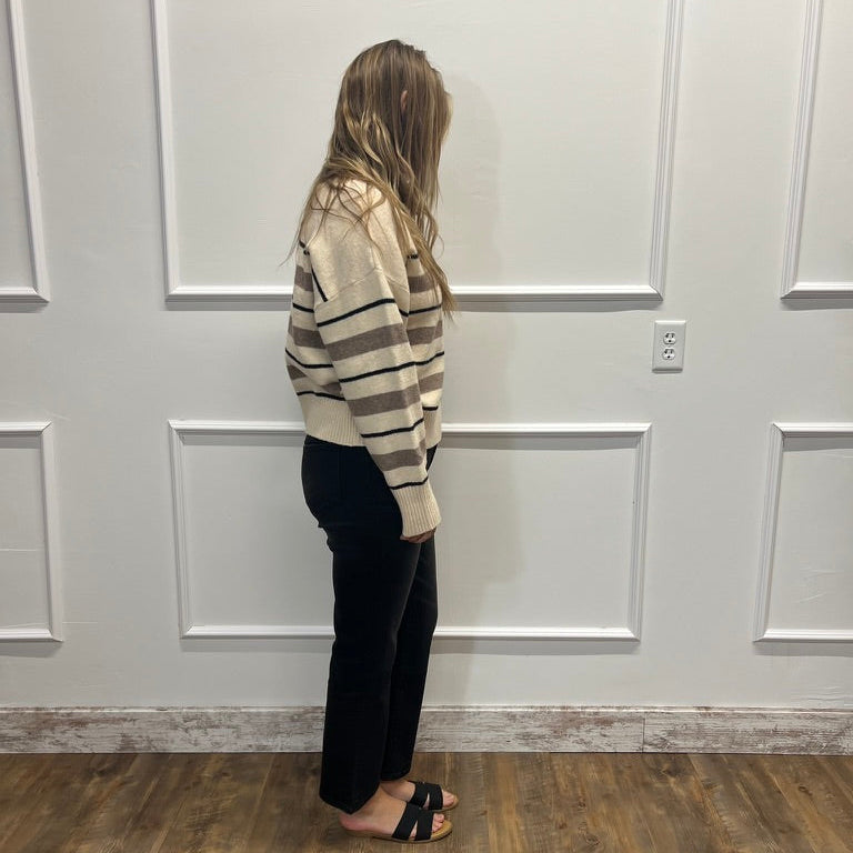 A woman stands in profile wearing a beige and brown striped knit sweater, black pants, and black slide sandals, standing on a wood-look floor against a white paneled wall.