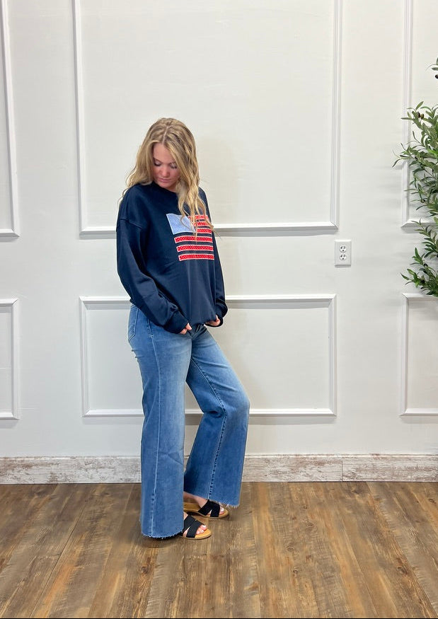 Woman wearing a navy sweatshirt with a colorful graphic and blue jeans standing in a room with white paneled walls and a plant.