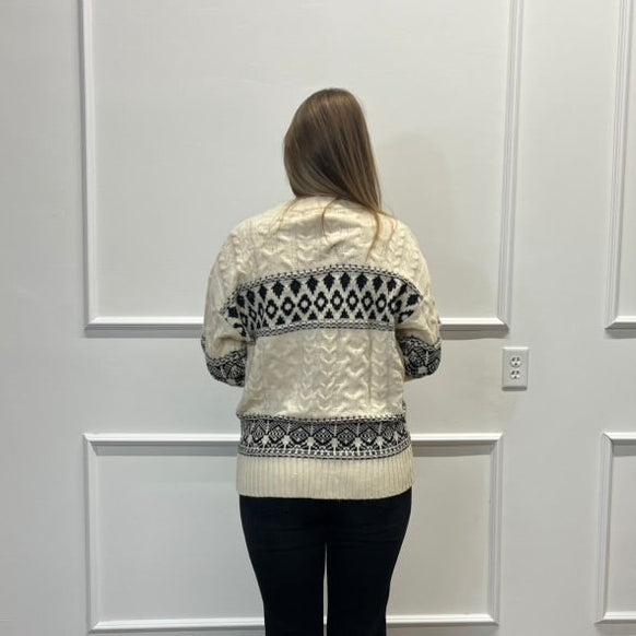 A woman viewed from behind wearing a cream cable-knit cardigan with a black geometric pattern across the shoulders, back, and hem, paired with black pants, standing in front of a white paneled wall on a wood floor.