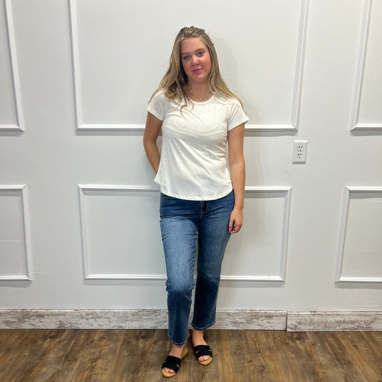 A young woman stands in front of a white paneled wall wearing a white short-sleeve t-shirt, blue jeans, and black slide sandals.