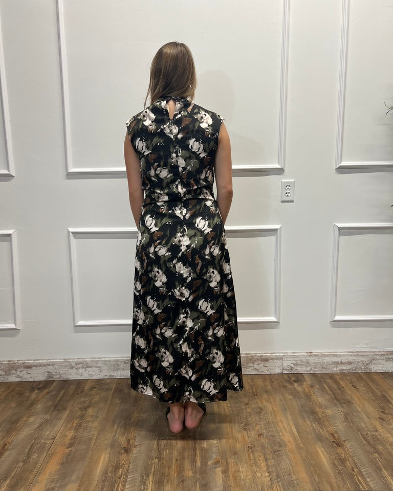A woman viewed from behind wearing a sleeveless black maxi dress with a white floral pattern, standing on a wood floor against a white paneled wall.