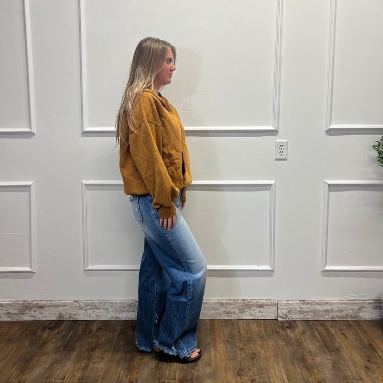 A woman stands in profile wearing a mustard yellow cropped jacket and wide-leg blue jeans, standing on a wood floor against a white paneled wall.