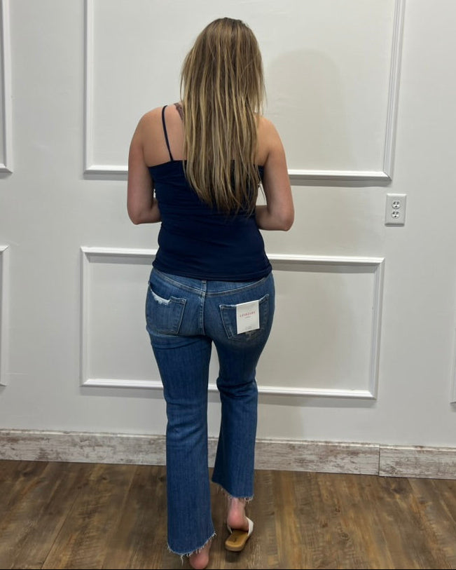 A woman viewed from behind wearing a dark navy blue camisole and blue jeans, standing in front of a white paneled wall on a wood floor.