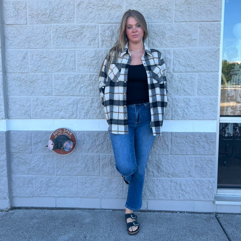 A woman stands against a light gray stone wall wearing a black and white buffalo plaid flannel shirt over a black tank top, blue jeans, and black sandals.