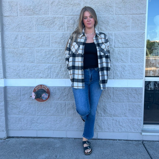 A woman stands against a light gray stone wall wearing a black and white buffalo plaid flannel shirt over a black tank top, blue jeans, and black sandals.