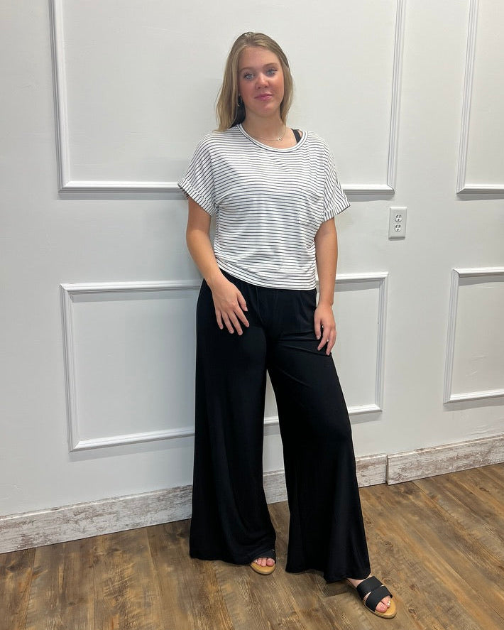 A woman modeling a short-sleeve striped t-shirt and black wide-leg pants, standing against a white paneled wall on a wood floor.