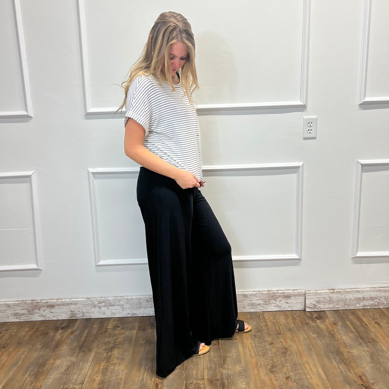 A woman stands in a room with white paneled walls and a wood floor, wearing a white short-sleeve t-shirt with thin black horizontal stripes and black wide-leg pants.