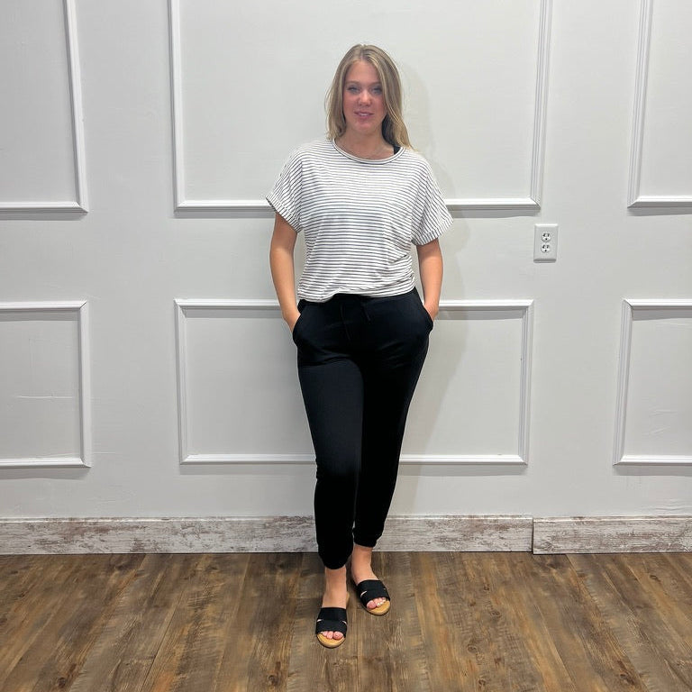 A woman stands in a room with white paneled walls and a wood floor, wearing a white short-sleeve striped t-shirt, black pants, and black slide sandals.