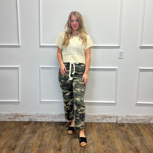 A woman stands in a room with white paneled walls and a wood floor, wearing a cream ribbed short-sleeve top, green camouflage drawstring pants, and black slide sandals.