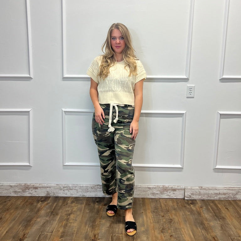 A woman stands in a room with white paneled walls and a wood floor, wearing a cream ribbed short-sleeve top, green camouflage drawstring pants, and black slide sandals.