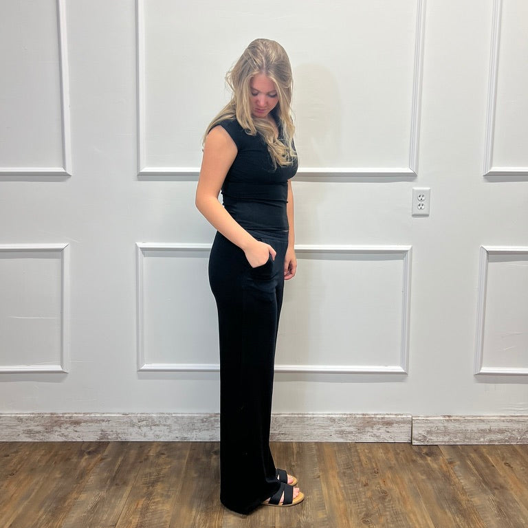 A woman modeling a sleeveless black maxi dress with a fitted bodice and flared legs, standing in front of a white paneled wall on a wood floor.