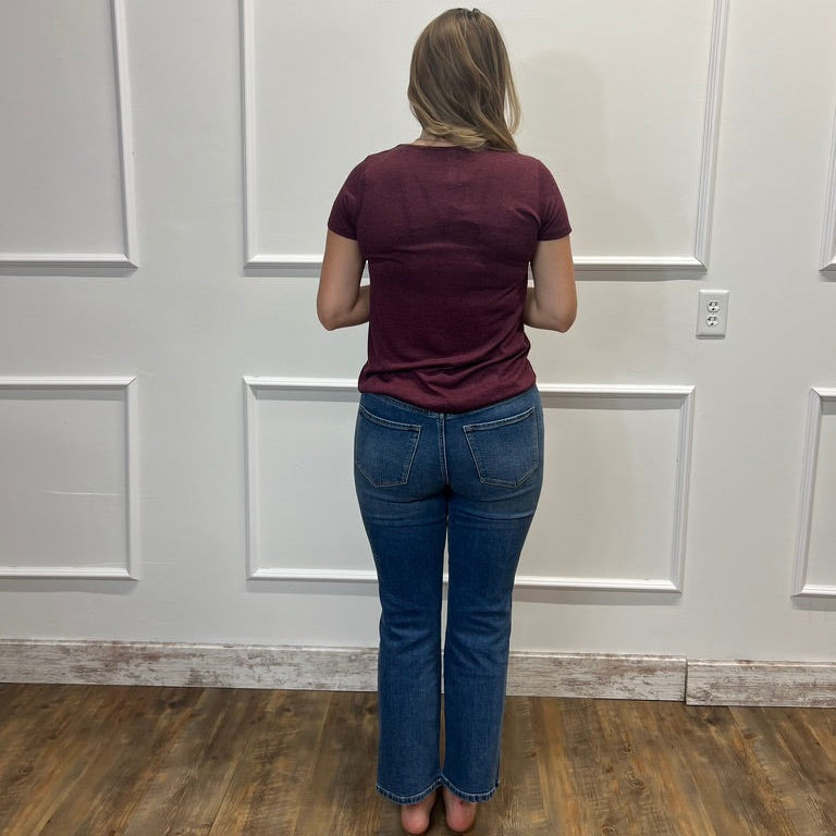 A woman viewed from behind wearing a maroon short-sleeve t-shirt and blue jeans, standing in a room with white paneled walls and a wood floor.